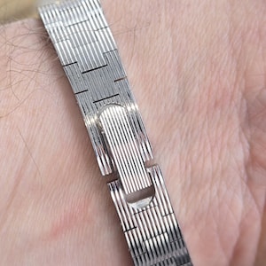 May include: A silver-colored watch band with a textured, ribbed design. The band is made of metal and has a clasp closure. The watch band is shown on a person's wrist.
