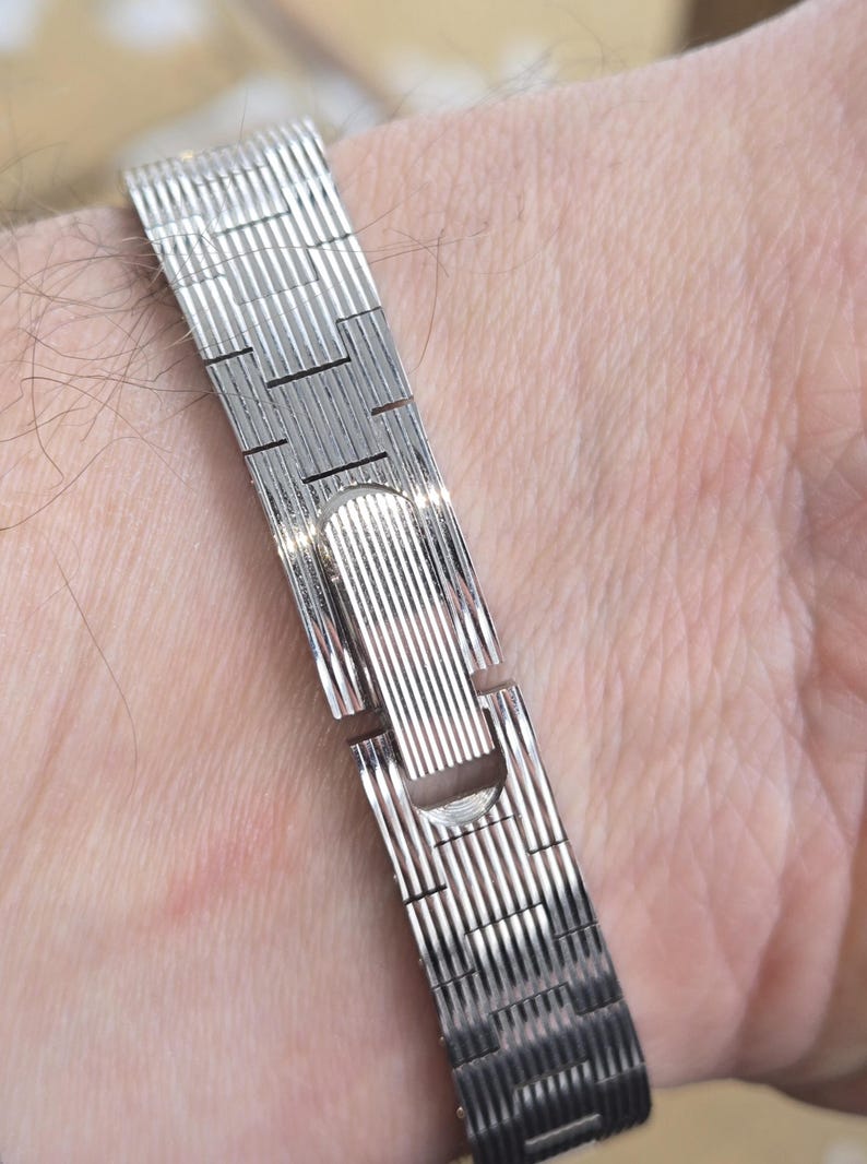 May include: A silver-colored watch band with a textured, ribbed design. The band is made of metal and has a clasp closure. The watch band is shown on a person's wrist.