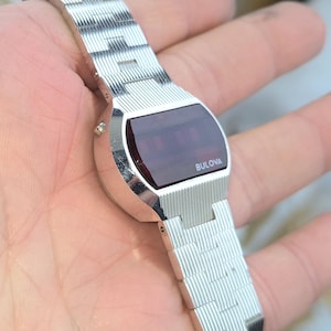 May include: A silver Bulova digital watch with a rectangular red display. The watch has a ridged metal band and the Bulova logo is visible on the watch face. The watch is held in a hand.