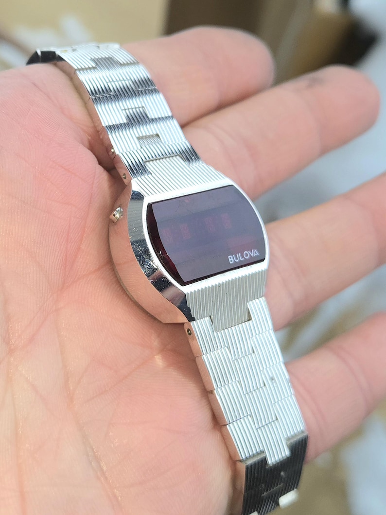 May include: A silver Bulova digital watch with a rectangular red display. The watch has a ridged metal band and the Bulova logo is visible on the watch face. The watch is held in a hand.