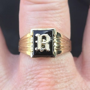 May include: A gold ring with a black rectangular centre stone. The stone features a raised, ornate letter "R" in a lighter gold tone. The ring has a ribbed design on the sides.