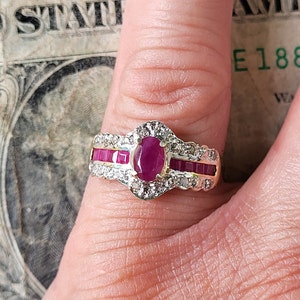 Könnte beinhalten: Ein goldener Ring mit einem großen ovalen Rubin, der von kleineren Diamanten und roten Rubinen umgeben ist. Der Ring ist in einer weißen Metallfassung gefasst.
