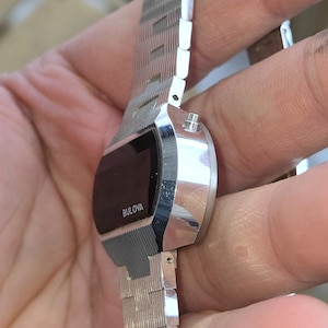 May include: A vintage Bulova digital watch with a silver-tone metal band and case. The watch face is black with the Bulova brand name in white. The band has a textured, geometric design.