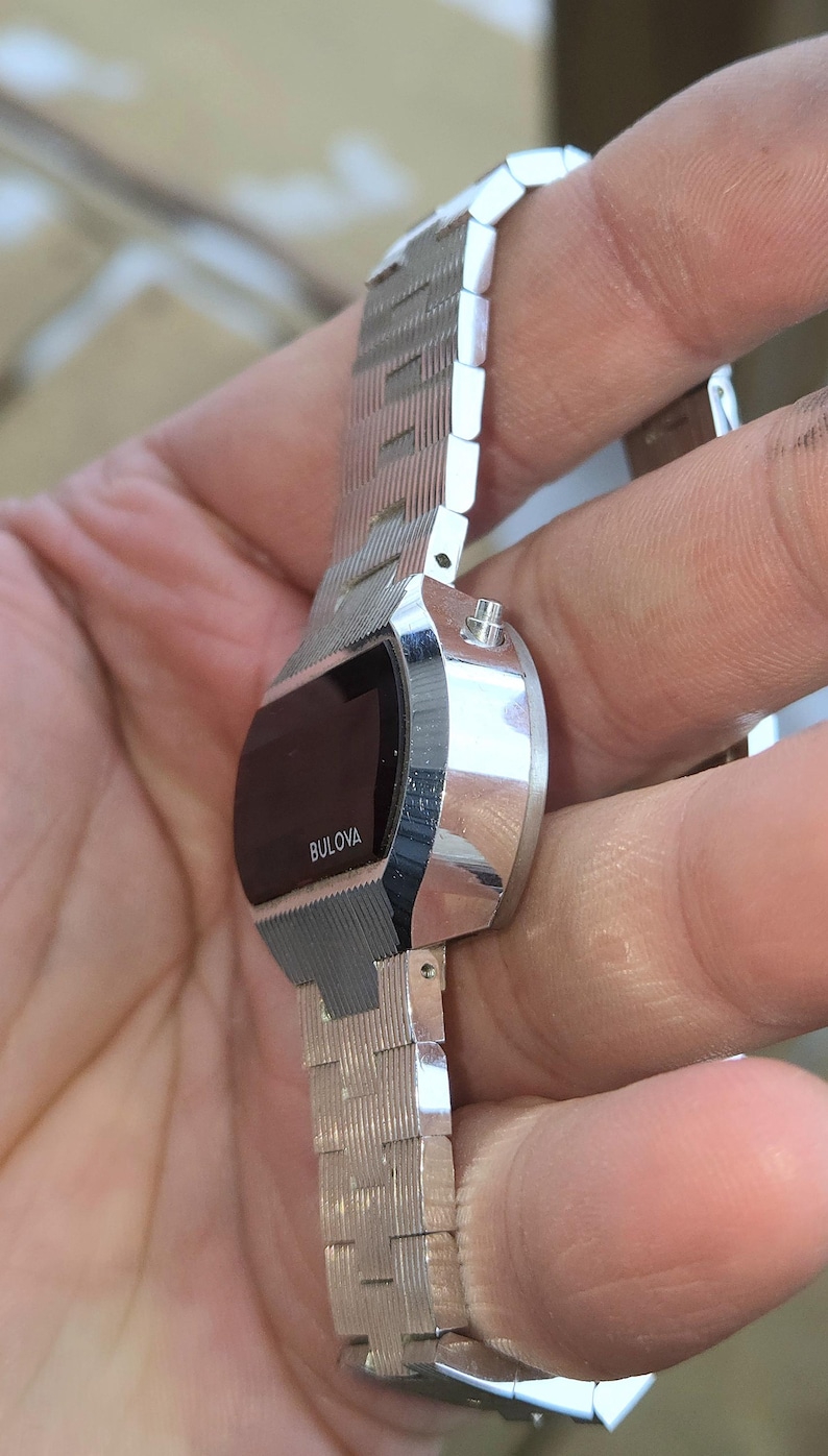 May include: A vintage Bulova digital watch with a silver-tone metal band and case. The watch face is black with the Bulova brand name in white. The band has a textured, geometric design.