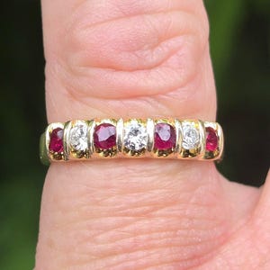 May include: A gold ring with alternating red and clear gemstones. The ring's design is classic, with the gemstones set in a row. The ring is displayed on a finger.