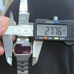 May include: A vintage Bulova digital watch with a silver band and a red display. The watch is being measured with a digital caliper, showing a measurement of 27.76 mm. The watch face has the Bulova logo.