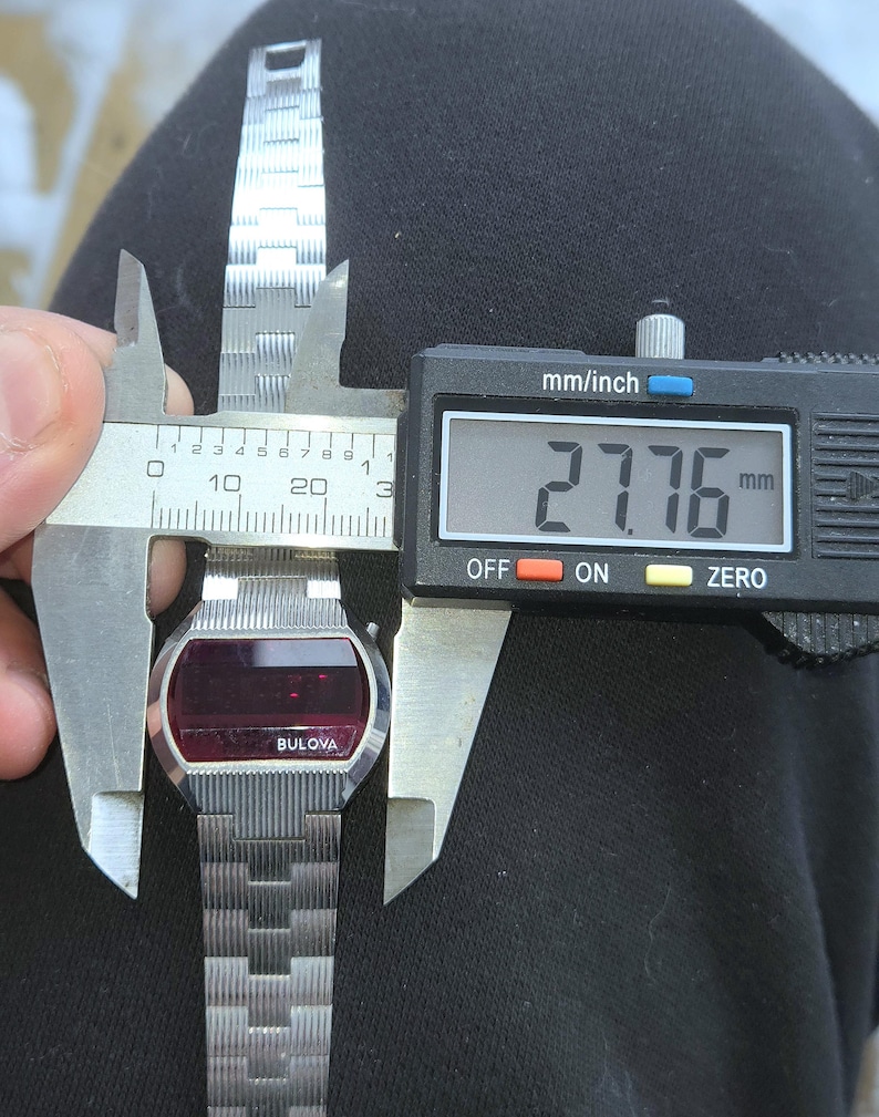 May include: A vintage Bulova digital watch with a silver band and a red display. The watch is being measured with a digital caliper, showing a measurement of 27.76 mm. The watch face has the Bulova logo.