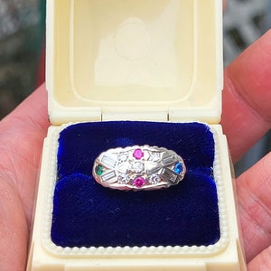 May include: A white ring box with a blue velvet interior, showcasing a silver ring with a row of small diamonds and a center stone surrounded by four colorful gemstones: green, pink, and blue.