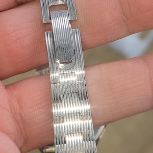May include: A silver-toned watch band with a textured, ribbed design. The band is made of connected rectangular segments and features a clasp. The watch band is held in a hand.
