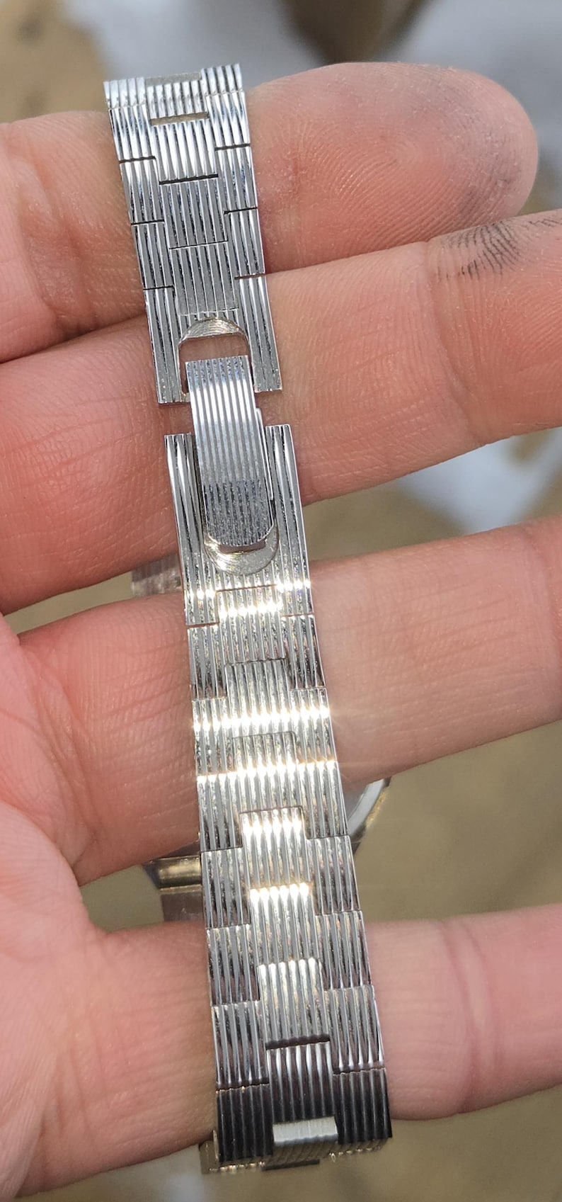 May include: A silver-toned watch band with a textured, ribbed design. The band is made of connected rectangular segments and features a clasp. The watch band is held in a hand.