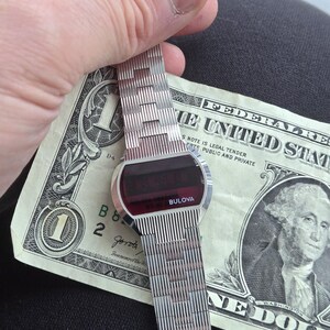 May include: A vintage Bulova digital watch with a silver band and a red digital display. The watch is held over a one-dollar bill. The watch face is oval-shaped and the band is made of small, rectangular links.