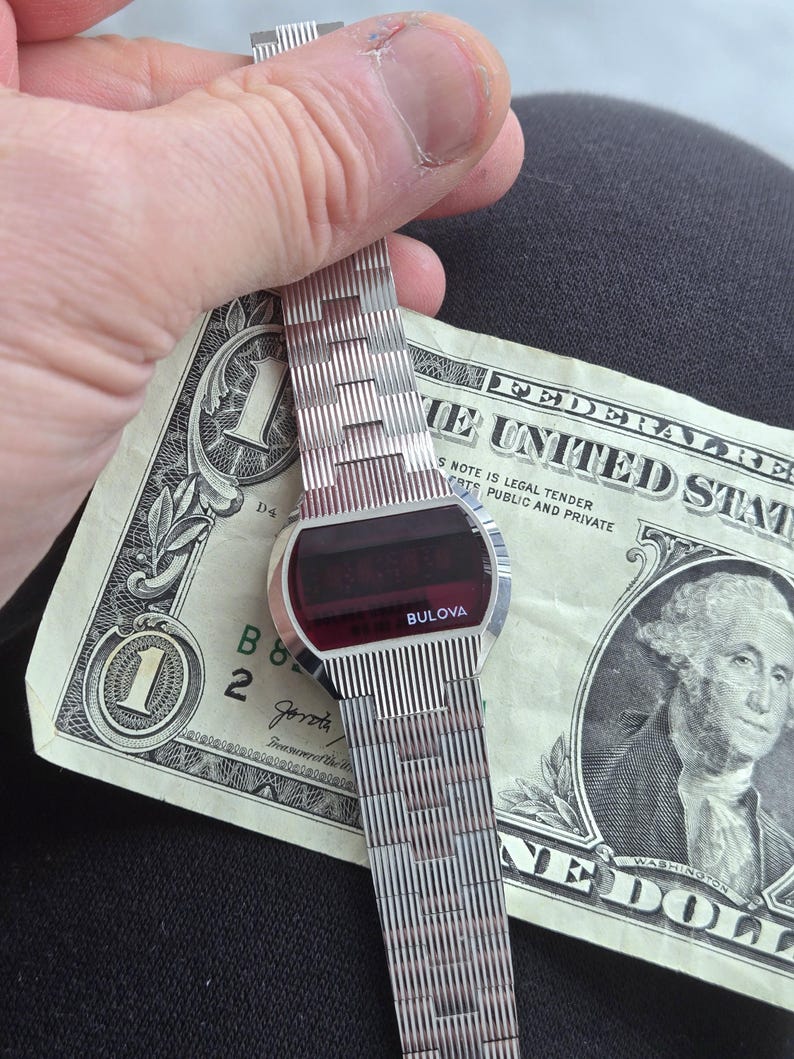 May include: A vintage Bulova digital watch with a silver band and a red digital display. The watch is held over a one-dollar bill. The watch face is oval-shaped and the band is made of small, rectangular links.