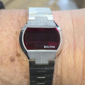 May include: A vintage Bulova digital watch with a silver-tone metal band and a rectangular red display. The watch face has the brand name "Bulova" in white. The watch is worn on a wrist.