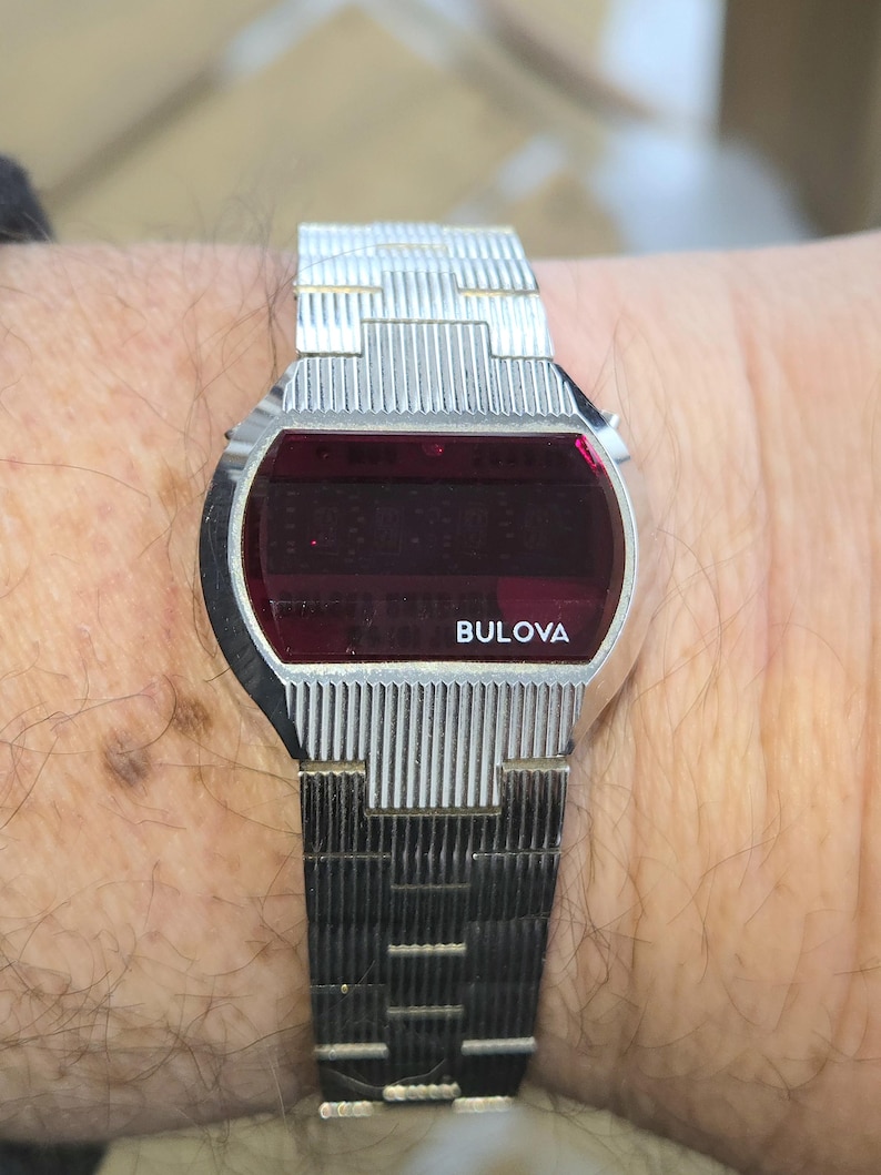 May include: A vintage Bulova digital watch with a silver-tone metal band and a rectangular red display. The watch face has the brand name "Bulova" in white. The watch is worn on a wrist.