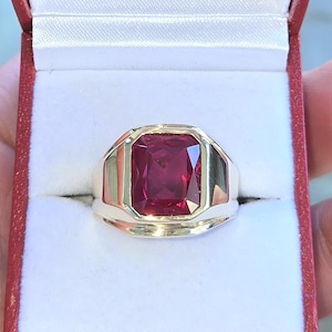 May include: A gold ring featuring a large, rectangular, deep red gemstone. The ring is set in a classic bezel setting, and is presented in a red jewellery box with a white interior. The ring's design is simple yet elegant.