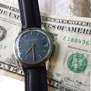 May include: A silver wristwatch with a blue face and black leather band. The watch face has silver hands, silver hour markers, and a date window at the 3 o'clock position. The watch is resting on a US dollar bill.