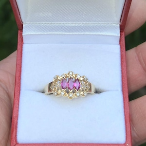 Könnte beinhalten: Ein goldener Ring mit drei rosa Edelsteinen in der Mitte, umgeben von kleineren weißen Steinen. Der Ring wird in einer roten Schmuckschatulle mit weißem Satineinsatz präsentiert. Das Design des Rings weist ein filigranes Muster auf.