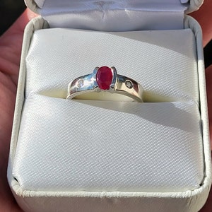 Può includere: Un anello d'argento con una pietra ovale rossa e due pietre bianche più piccole. L'anello è in una scatola per anelli bianca.