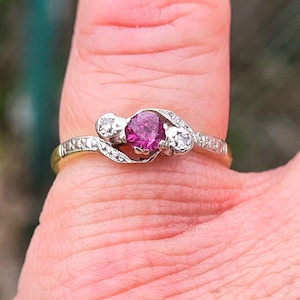 Könnte beinhalten: Ein zarter Ring mit einem rosafarbenen Edelstein, der von kleineren weißen Edelsteinen umgeben ist. Der Ring ist in einer verdrehten Bandfassung gefasst.
