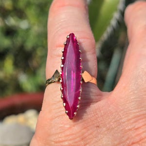 May include: A gold-toned ring featuring a large, elongated, magenta-colored gemstone. The gemstone is faceted and surrounded by a gold-colored setting with small, decorative prongs. The ring's band has a unique, geometric design.
