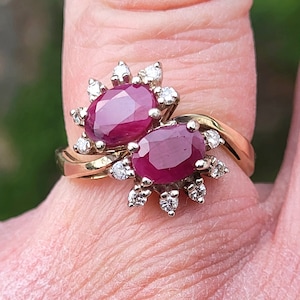 Könnte beinhalten: Ein goldener Ring mit zwei ovalen Rubin-Edelsteinen, die von kleineren Diamanten umgeben sind.