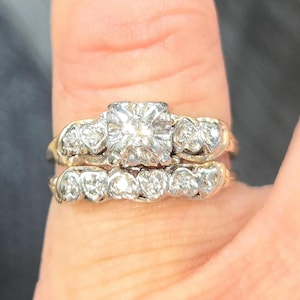 May include: A two-piece wedding ring set. The engagement ring features a large, square-cut diamond center stone, flanked by smaller diamonds. The matching band has multiple small diamonds. Both rings have a gold band.