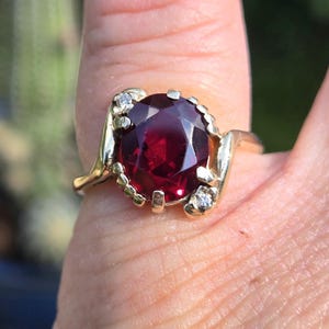 May include: A gold ring featuring a large, oval-shaped, red gemstone surrounded by smaller, clear gemstones.