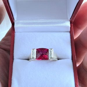 May include: A red gemstone ring with a square-shaped stone set in a silver band. The ring is surrounded by smaller white gemstones.