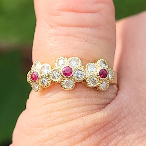 Könnte beinhalten: Ein goldener Ring mit drei blütenförmigen Fassungen, jede mit einem kleinen roten Edelstein in der Mitte und umgeben von kleineren weißen Edelsteinen.