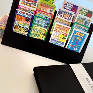 Puede incluir: Un soporte organizador de tela negra que contiene múltiples tarjetas de bingo coloridas con varios títulos como Rodeo, Triple Emeralds y Twice the Fire. El soporte cuenta con ranuras escalonadas para mostrar las tarjetas y un estuche de tela negra con una etiqueta blanca.