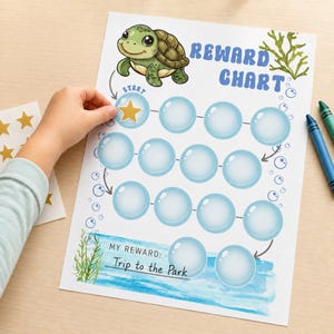 May include: A reward chart printed on white paper featuring a green cartoon turtle, blue bubble-shaped spaces for stickers, and the text "REWARD CHART" and "MY REWARD: Trip to the Park." A gold star sticker is being placed in the first bubble.