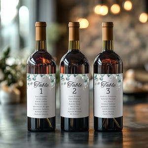 May include: Three wine bottles with brown caps and labels featuring eucalyptus and guest names. Each label reads "Table" followed by a number, and lists names. The bottles are filled with red wine.