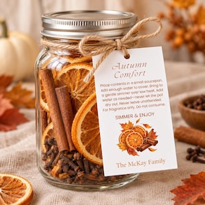 Peut inclure: Un bocal en verre transparent rempli de tranches d'orange séchées, de bâtons de cannelle et de clous de girofle. Une ficelle de jute maintient une étiquette rectangulaire blanche avec le texte Autumn Comfort et des illustrations.