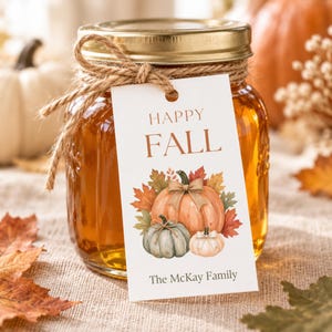 May include: A glass jar filled with amber liquid, topped with a gold lid and tied with twine. A white rectangular tag hangs from the twine, featuring the text HAPPY FALL, an illustration of three pumpkins with autumn leaves, and The McKay Family.