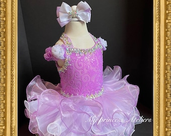 May include: A lavender and white layered tulle dress with a beaded bodice and a matching lavender bow headband. The dress is designed for a young girl and features a ruffled skirt.