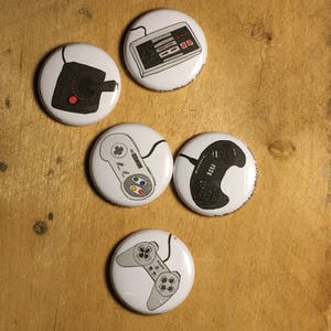 Puede incluir: Una colección de pines blancos con diseños de mandos de videojuegos clásicos. Los pines muestran mandos de Atari, Nintendo, Sega y Sony, con gráficos en negro y gris. Los botones están dispuestos sobre una superficie de madera.