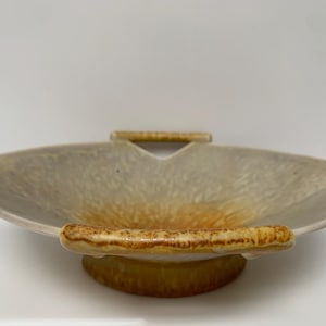 May include: A ceramic serving bowl with a distinctive, leaf-like form. The bowl has a light beige interior with orange and brown gradients. It features two handles and a circular base, all in a similar colour palette.