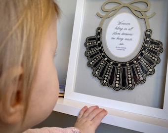 May include: A framed print of a black and white collar with rhinestones. The print includes the quote "Women belong in all places decisions are being made." and the name "Ruth Bader Ginsburg".