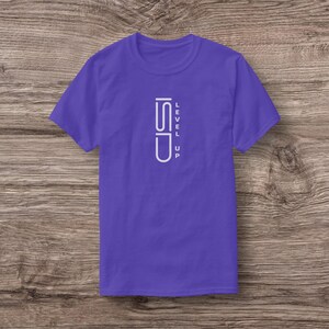 Puede incluir: Una camiseta morada con las palabras "LEVEL UP" en blanco, alineadas verticalmente en la parte delantera. El diseño presenta un gráfico estilizado. La camiseta está hecha de un material suave y tiene mangas cortas.