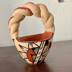 May include: A small, hand-crafted pottery basket with a twisted handle. The basket is decorated with a geometric pattern in red, black, and white.