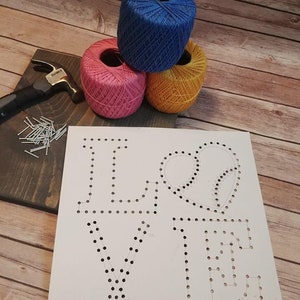 May include: A white card with the word "LOVE" written in black with a heart in the middle. The card has holes punched in the shape of the letters and heart. There are three balls of yarn in the background: blue, pink, and yellow. There are also nails and a hammer on a wooden surface.