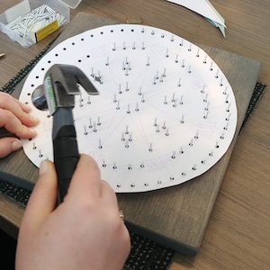 May include: A person hammering nails into a circular wooden board with a compass rose design and the letters N, NE, E, SE, S, SW, W, and NW printed around the edge.