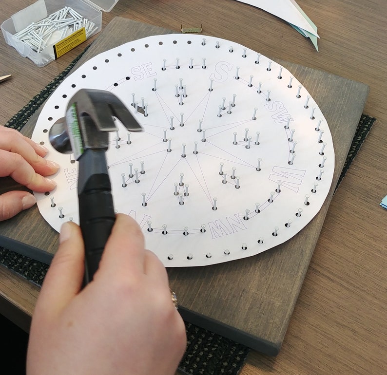 May include: A person hammering nails into a circular wooden board with a compass rose design and the letters N, NE, E, SE, S, SW, W, and NW printed around the edge.