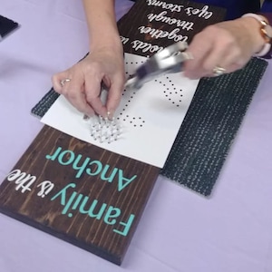 May include: A person using a tool to hammer nails into a white piece of paper with a pre-printed anchor design. The paper is placed on top of a wooden sign that reads "Family is the Anchor".