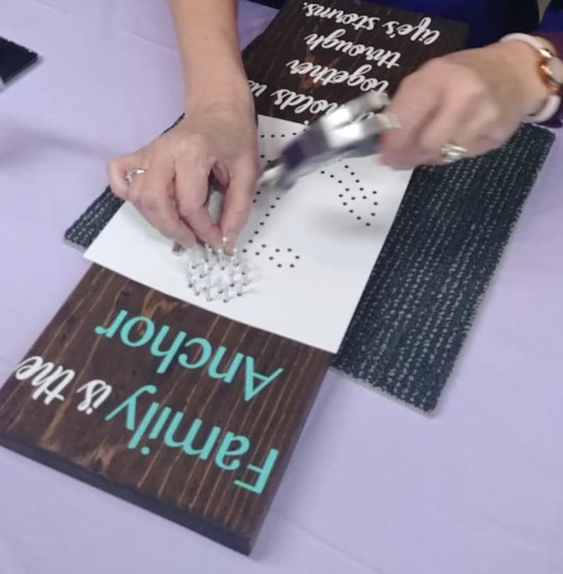 May include: A person using a tool to hammer nails into a white piece of paper with a pre-printed anchor design. The paper is placed on top of a wooden sign that reads "Family is the Anchor".