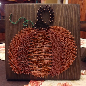 May include: A string art pumpkin with an orange and green color scheme. The pumpkin is made of string wrapped around nails hammered into a wooden board.