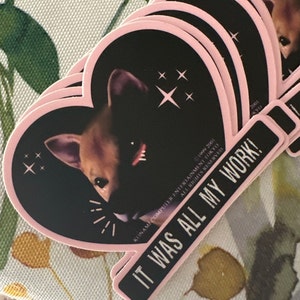 May include: A stack of heart-shaped stickers with a black background and a pink border. The stickers feature a dog's face and starburst designs. A rectangular sticker with the text "IT WAS ALL MY WORK!" is also visible.