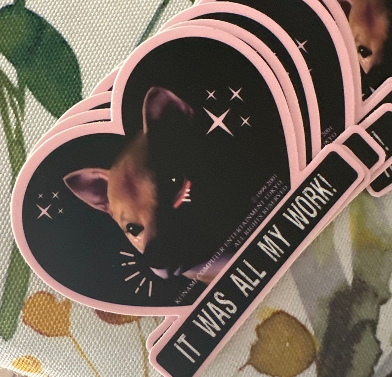 May include: A stack of heart-shaped stickers with a black background and a pink border. The stickers feature a dog's face and starburst designs. A rectangular sticker with the text "IT WAS ALL MY WORK!" is also visible.
