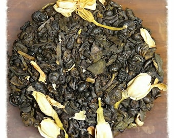 May include: A close-up of a blend of green tea leaves and dried jasmine flowers. The tea is a dark green color and the flowers are a pale yellow.