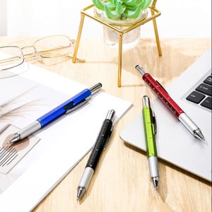 Puede incluir: Cuatro bolígrafos multiherramienta en azul, negro, rojo y verde con detalles plateados. Cada bolígrafo incluye una regla, un nivel y una punta de lápiz táctil. Los bolígrafos están colocados sobre una superficie de madera clara, con un portátil y un pequeño soporte dorado para plantas en el fondo.